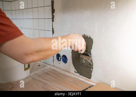 Worker's hand applying adhesive mortar on a white wall using a trowel, installing ceramic tiles in a kitchen or bathroom as part of a home renovation Stock Photo