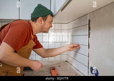 Professional tiler carefully installing new white ceramic tiles on a kitchen wall, demonstrating precision and expertise during home improvement and r Stock Photo