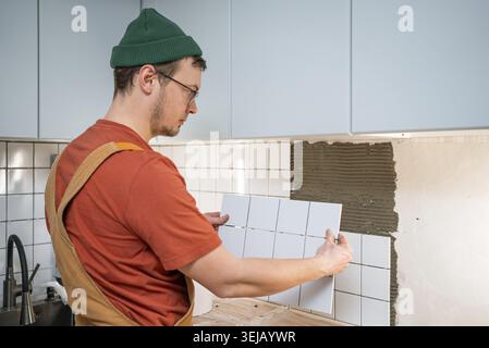 Man wearing a green hat and orange shirt carefully installing new white ceramic tiles on the kitchen wall, renovating the backsplash with adhesive dur Stock Photo