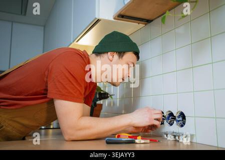 Electrician installing new kitchen outlet on a tiled backsplash after renovation, connecting electrical wires, providing home improvement and professi Stock Photo