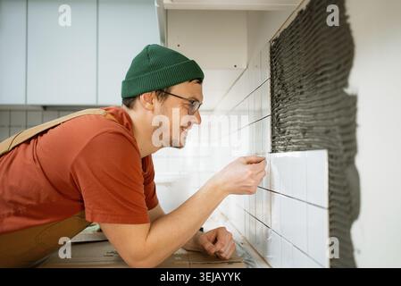Man smiling while accurately laying white ceramic tiles on a kitchen wall, focusing on each piece to complete a new backsplash during home improvement Stock Photo