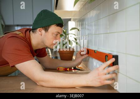 Man wearing a green hat concentrating on checking the horizontal level of newly installed electrical outlets on a tiled kitchen wall using a spirit le Stock Photo