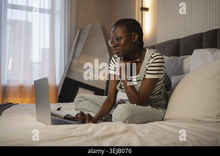 Young african american woman wearing glasses is sitting comfortably on her bed. Deeply engrossed in her work on a laptop. With a notebook beside her. Stock Photo