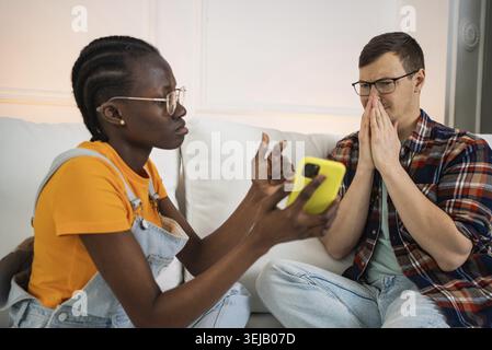 Young woman showing shocking news or photos from social media on smartphone to stressed man sitting on sofa at home, he is covering his mouth with his Stock Photo