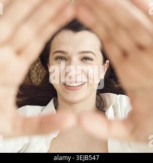 Caucasian young female smiling through triangle formed by hands, close-up portrait with focus on face and eyes Stock Photo