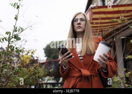 Seriously, a young beautiful woman walks around the city with a cup of coffee and a phone in her hands Stock Photo