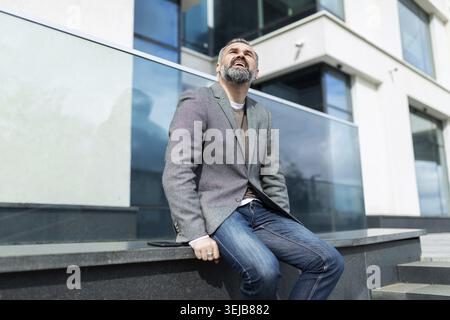Elderly man businessman with a gray beard sits near the office building and looks up Stock Photo