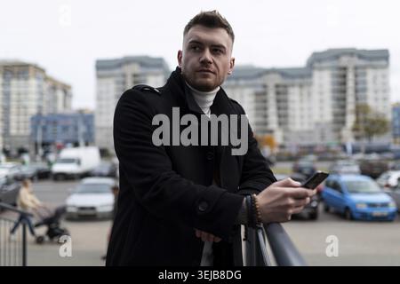 Portrait of a brutal young businessman in a stylish black coat with a phone in his hands on the background of the city Stock Photo