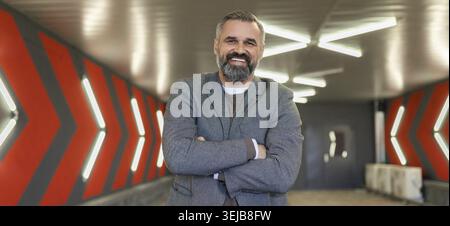 A gray-haired fifty-year-old man businessman in an underground parking lot stands with his arms folded across his chest Stock Photo