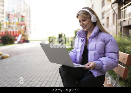 Stylish modern woman working on laptop with headphones outside Stock Photo