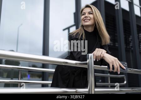Business successful middle-aged blonde woman with a smile looks into the distance against the backdrop of an office building Stock Photo