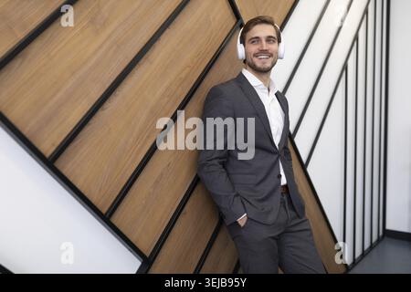 Young businessman in headphones in a business suit listening to music leaning against the wall in the office Stock Photo