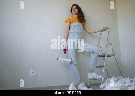 Female painter is taking a break while holding a paint roller and leaning on a ladder during a home renovation project, with old wallpaper scraps on t Stock Photo
