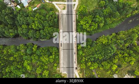 An overhead drone shot captures a straight asphalt road on a bridge spanning a calm river through dense woodland, showcasing nature's tranquility Stock Photo