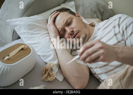 Sick man with fever symptoms is lying in bed and checking his body temperature with a thermometer, suffering from seasonal flu or other virus Stock Photo