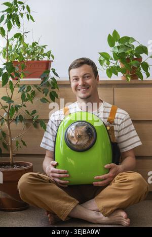 Young man sitting cross-legged on the floor, smiling and holding a cat inside a space backpack carrier, surrounded by houseplants in his modern apartm Stock Photo