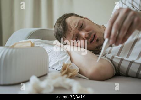 Caucasian man suffering from cold or flu is lying in bed at home, checking his body temperature with a thermometer, with tissues and tissue box visibl Stock Photo
