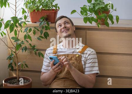 Happy male gardener sitting on the floor in brown overalls, smiling while using a smartphone to read plant care instructions and learning about nurtur Stock Photo