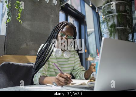 Freelancer african american woman taking notes while working on laptop in trendy cafe and sipping drink, multitasking with headphones and blue drink, Stock Photo