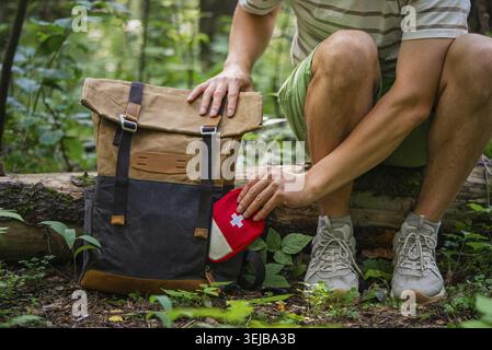 Man placing a first aid kit into his hiking backpack while resting in a forest, preparing for outdoor adventure Stock Photo