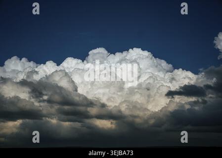 Bright white cumulus clouds are forming above a dark landscape, creating a dramatic contrast against the deep blue sky, hinting at an approaching stor Stock Photo