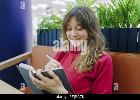 Young student smiling while taking notes and preparing for an upcoming exam, sitting comfortably at a table in a modern cafe, enjoying a relaxed atmos Stock Photo