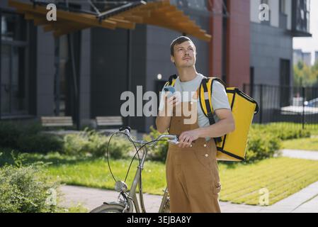 Male bike courier wearing overalls and carrying a yellow delivery backpack, using a smartphone to find a delivery location in the city, representing u Stock Photo