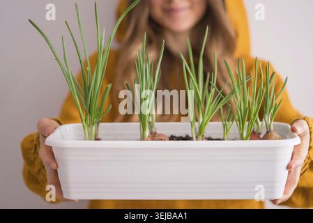Smiling woman in vibrant yellow sweater holds container of freshly sprouted green onions, showcasing her success in home gardening Stock Photo