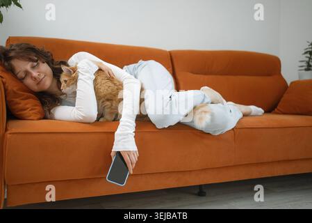 Exhausted woman sleeping on an orange sofa, cuddling her ginger cat while her phone slips from her hand, illustrating the concepts of rest, relaxation Stock Photo