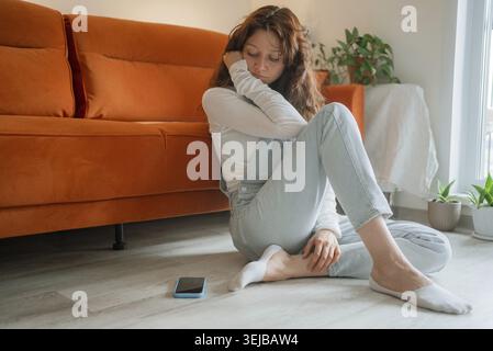 Sad woman sitting on the floor beside the sofa, lost in deep thoughts, grappling with feelings of loneliness and isolation in the comfort of her home Stock Photo