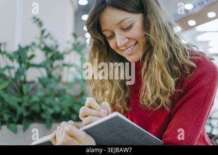 Young woman with long blond curly hair and red turtleneck sweater, smiling while writing on a notebook in a public space with plants in the background Stock Photo