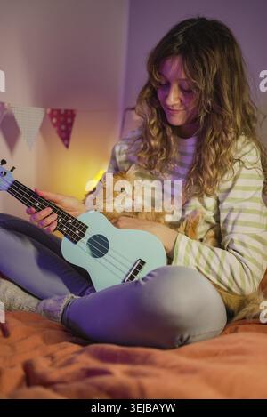 Young woman sitting cross-legged on a bed, strumming a light blue ukulele while gently stroking a ginger cat in her lap, enjoying a cozy indoor hobby Stock Photo