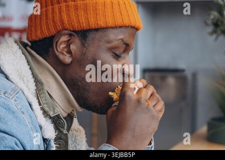 Young african american man wearing bright orange winter beanie, biting into juicy burger inside casual fast food restaurant, side profile capturing wi Stock Photo