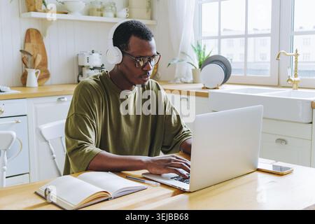 Young african american male programmer wearing headphones and glasses is concentrating on his work on a laptop in a bright home kitchen, demonstrating Stock Photo