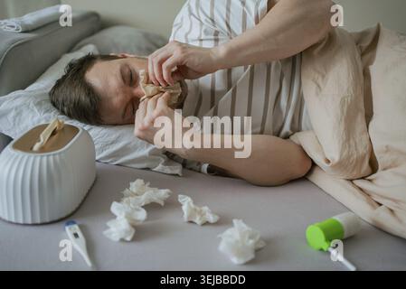 Sick man lying in bed, blowing his nose with tissues, surrounded by a thermometer, nasal spray, and a tissue box on the sheets, illustrating the strug Stock Photo