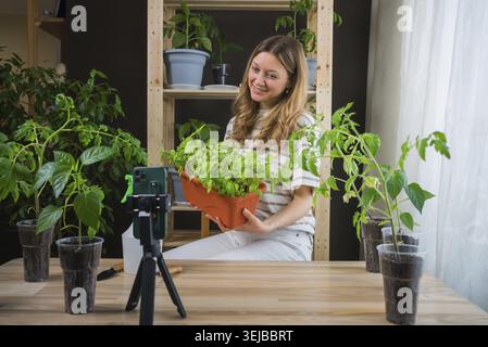 Smiling woman holding a pot filled with aromatic herbs while recording a video blog about home gardening, using her smartphone set on a tripod to capt Stock Photo