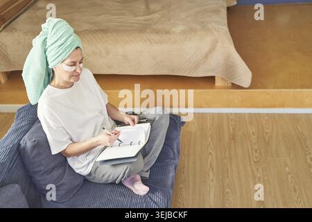 Caucasian mature female sitting on floor writing in notebook at home with under eye patches during calm selfcare reflection routine Stock Photo