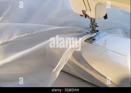 A sewing machine is in use, with a white fabric being sewn. The machine is in the foreground, and the fabric is in the background. Closeup Stock Photo