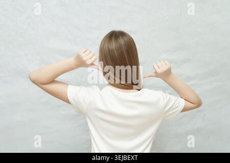 Rear view of young caucasian female in white t-shirt pointing thumbs at herself against light background Stock Photo