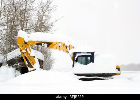 Spirit River, Alberta, Canada - December 26, 2025: yellow CAT excavator in the farm yard under heavy snow after snowfall. Little old barn and trees in Stock Photo