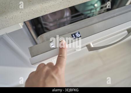 Female hand pressing button on modern dishwasher control panel in kitchen setting Stock Photo