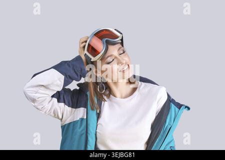 Young caucasian female wearing ski jacket and goggles in relaxed pose on gray background Stock Photo