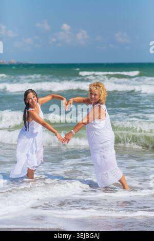 Two happy mature women making heart shape with arms in ocean waves enjoying friendship love happiness and carefree summer vacation lifestyle Stock Photo