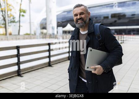 Mature man with laptop and backpack hurrying to work smiling looking at camera Stock Photo