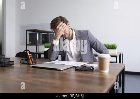 Tired upset businessman sits in front of the desktop in the office covering his face with his palm Stock Photo