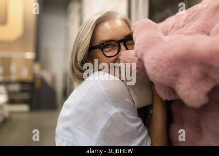 A woman with grey hair and glasses is happily embracing a soft pink blanket in a home decor store, surrounded by shelves of various textiles, enjoying Stock Photo