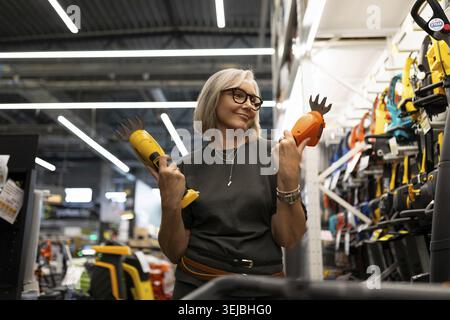 A woman with glasses smiles while holding two gardening tools, contemplating her choice in a home improvement store. Bright lighting highlights the pr Stock Photo