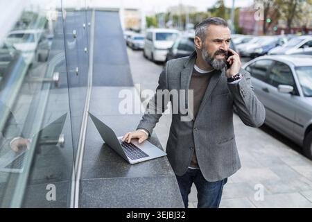 Mature business man in a jacket with a laptop talking on the phone outside Stock Photo