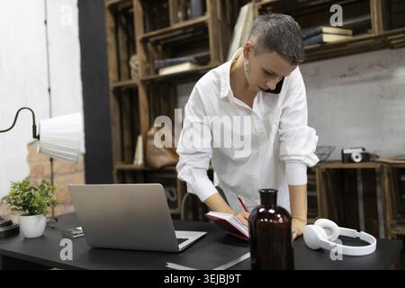 A woman with a short haircut works in the office talking on the phone making notes in a notebook Stock Photo
