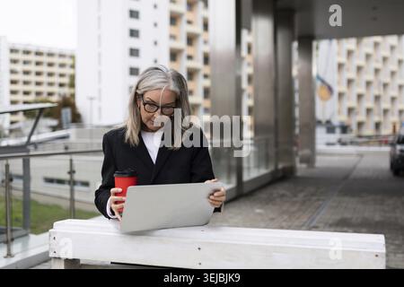 Senior businesswoman working on a laptop on the terrace of an office building with a cup of coffee in her hands Stock Photo